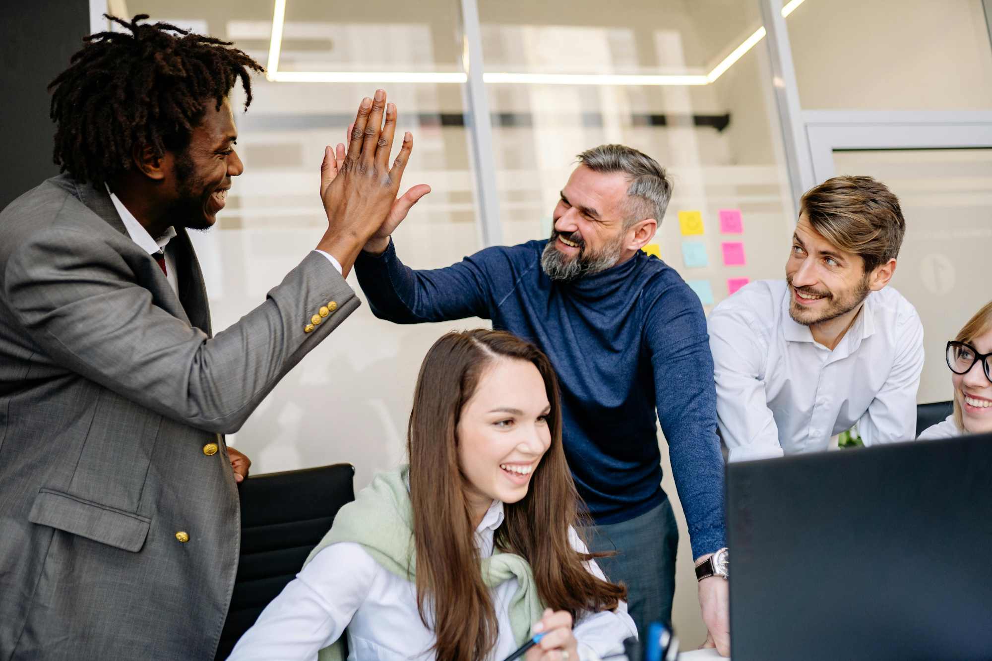 team members in a modern office smiling and high-fiving while gathered around a computer, celebrating a successful collaboration